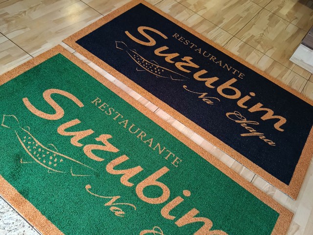Dois tapetes Cleankap do Restaurante Surubim na Chapa, um azul-marinho e um verde, ambos com borda laranja e logotipo dourado