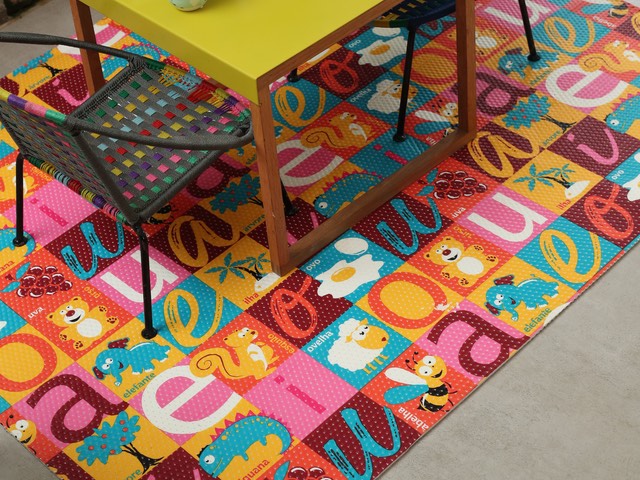 Tapete infantil com estampa de vogais colorida, cada letra num quadrado com animais, sob uma mesinha de brincar
